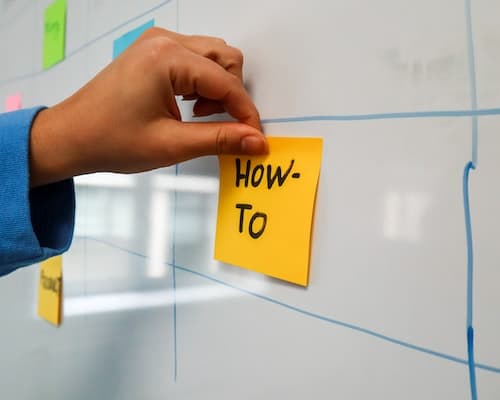 Person sticking a post it note on board that says How-to.