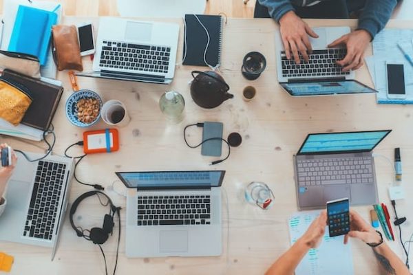 Big desk with one person writing on laptop, another checking mobile phone, different laptop open, phones, pens, markers, ear phone, etc.