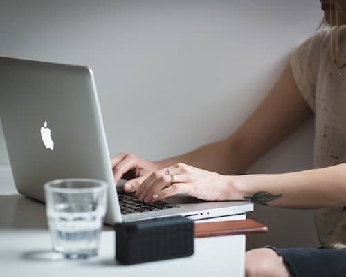 Woman typing on MacBook on desktop with class of water next to her.